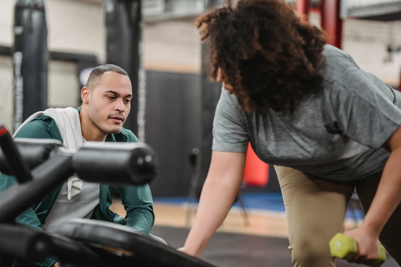 gallery-6 Experienced instructor watching anonymous African American female lifting dumbbell on blurred background of gym