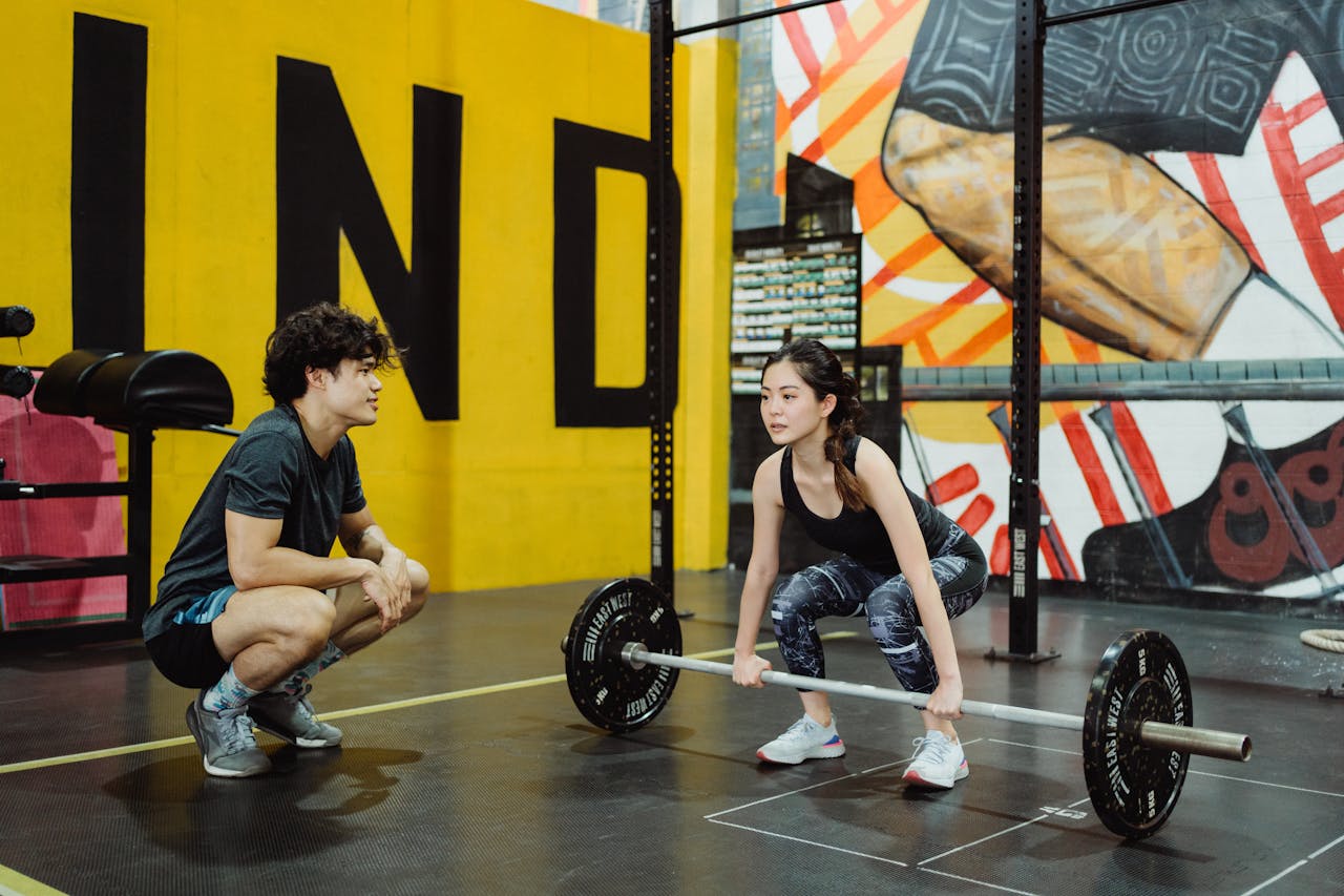 hero-img-02 A young woman lifting weights guided by a trainer in a colorful gym setting.